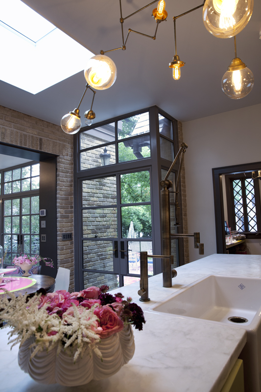 A kitchen with custom copper lights, a white marble countertop, a Rohl farmers sink, steel and glass doors and windows, a brick wall, and pink flowers.